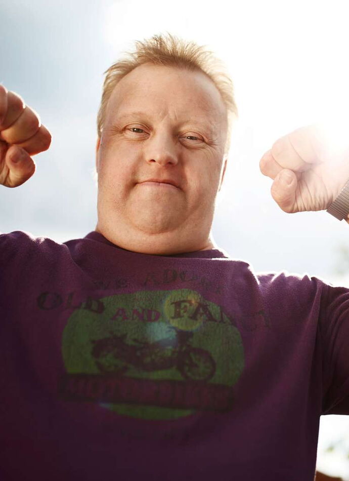 Ein Mann mit Down-Syndrom steht im Freien und reckt beide Fäuste triumphierend in die Höhe. Er trägt ein lila T-Shirt mit Motorrad-Aufdruck. Das Foto ist von unten aufgenommen – im Hintergrund Himmel und grüne Bäume, Sonnenlicht bricht hinter seinem Kopf