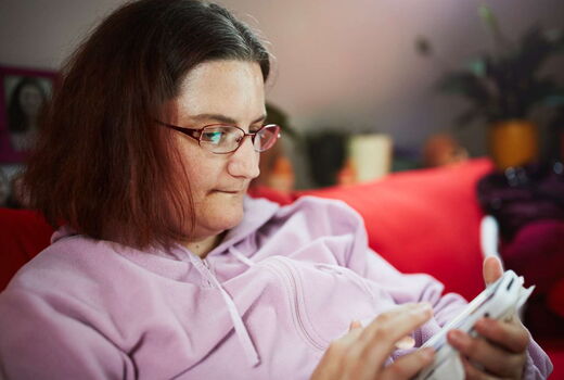 Eine Frau mit Brille sitzt auf einem roten Sofa und schaut auf ein Smartphone in ihren Händen. Sie trägt einen hellrosa Kapuzenpullover und wirkt konzentriert. Im Hintergrund sind unscharf Pflanzen und Gegenstände in einem Wohnraum zu sehen.