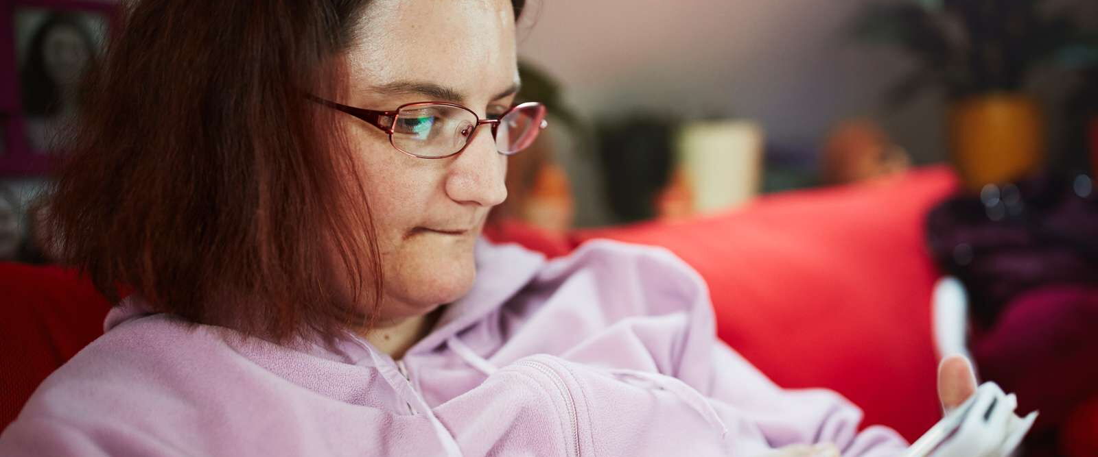 Eine Frau mit Brille sitzt auf einem roten Sofa und schaut auf ein Smartphone in ihren Händen. Sie trägt einen hellrosa Kapuzenpullover und wirkt konzentriert. Im Hintergrund sind unscharf Pflanzen und Gegenstände in einem Wohnraum zu sehen.