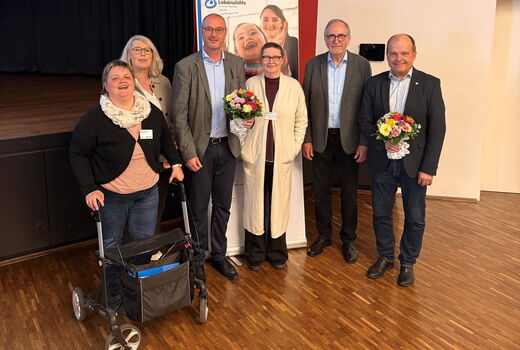 Sechs Personen posieren lächelnd für ein Gruppenfoto in einem Veranstaltungssaal. Im Hintergrund ist ein Roll-up-Banner der Lebenshilfe NRW zu sehen. Zwei der abgebildeten Personen halten bunte Blumensträuße in den Händen.