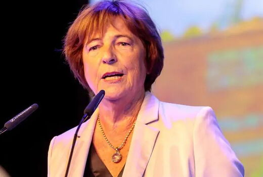 Eine Frau steht hinter einem Rednerpult und spricht in zwei Mikrofone. Sie trägt einen hellen Blazer, ein dunkles Oberteil und eine Kette.