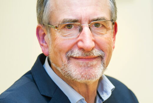 Ein Mann mit grauen Haaren, Bart und Brille schaut in die Kamera. er trägt ein Blaues Sako.