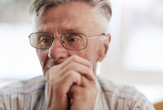 Nahaufnahme eines älteren Mannes mit Brille. Er hält die Hände vor dem Gesicht zusammen und schaut konzentriert nach unten. Im Hintergrund fällt helles Tageslicht durch ein Fenster.