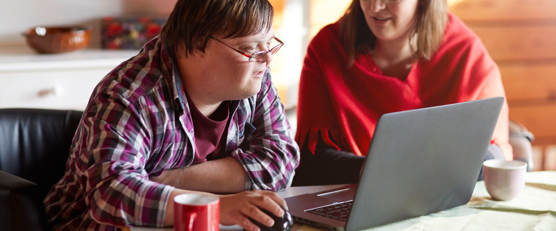 Ein junger Mann mit Down-Syndrom und eine Frau sitzen gemeinsam an einem Tisch und schauen auf einen Laptop-Bildschirm.