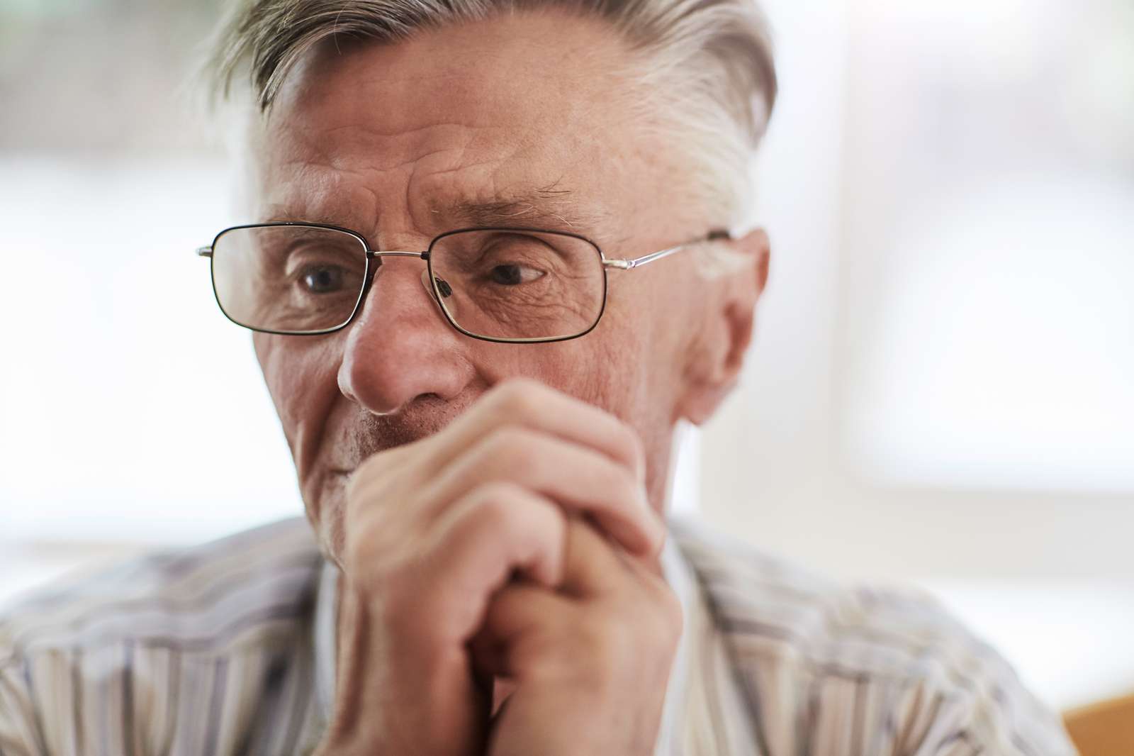 Nahaufnahme eines älteren Mannes mit Brille. Er hält die Hände vor dem Gesicht zusammen und schaut konzentriert nach unten. Im Hintergrund fällt helles Tageslicht durch ein Fenster.