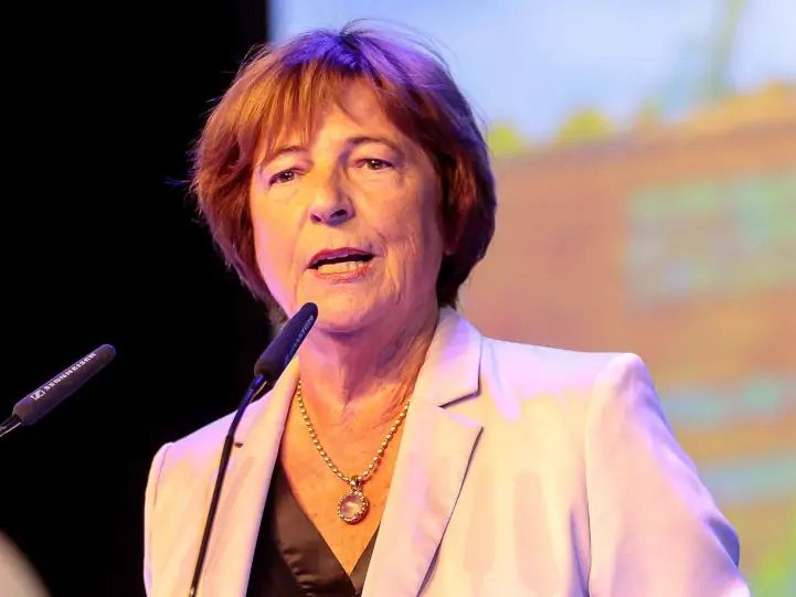 Eine Frau steht hinter einem Rednerpult und spricht in zwei Mikrofone. Sie trägt einen hellen Blazer, ein dunkles Oberteil und eine Kette.